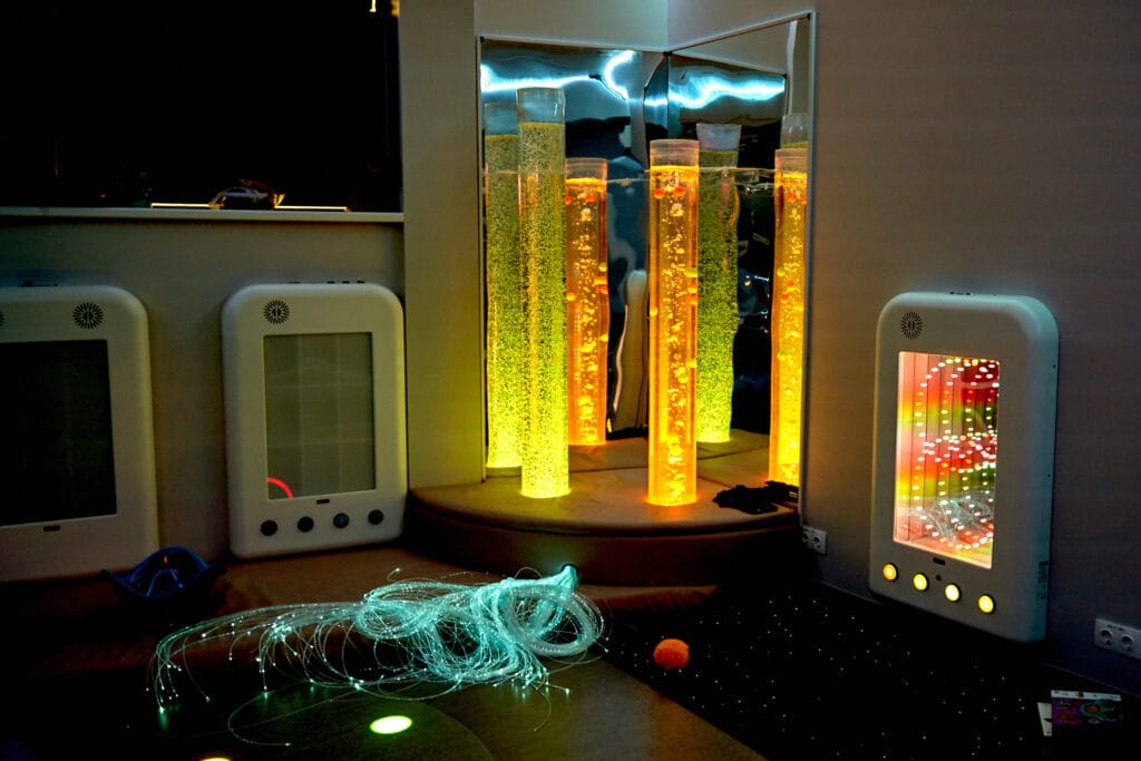 A dimly lit multi-sensory room containing multi-coloured bubble tube lamp, fairy lights, fluffy stress balls and other sensory materials.