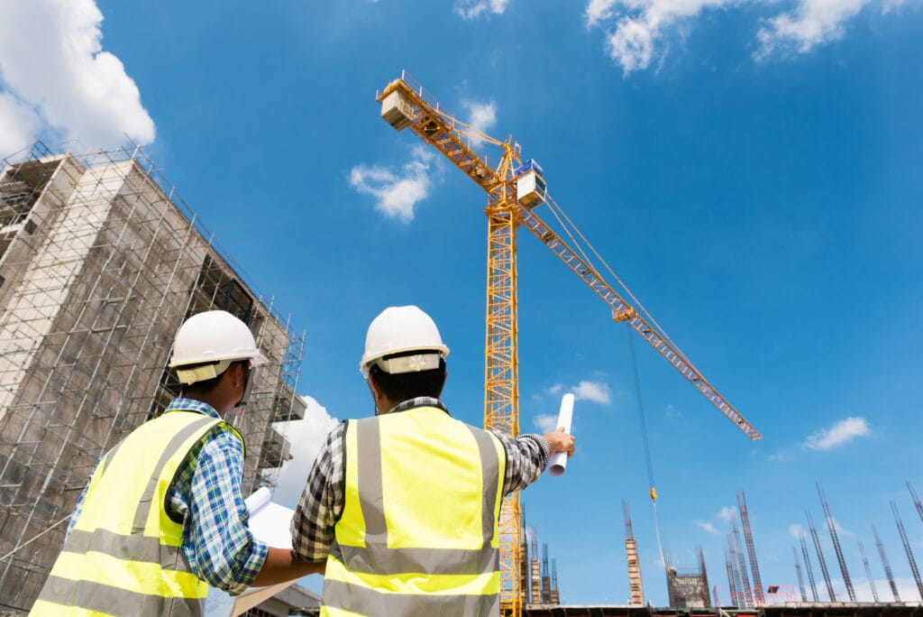 Two male engineers wearing hi-vis jackets and white hard hats have a discussion on a building site on a sunny day.