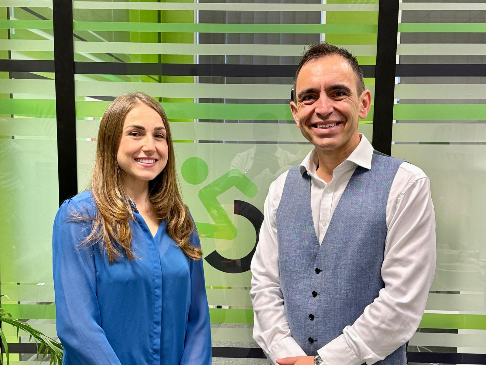 The image shows two individuals (Steven Mifsud and Jude Mifsud) standing indoors in front of a glass wall with a decorative design. The glass wall features horizontal frosted stripes and a large green accessibility symbol, which resembles a stylized person in a wheelchair. Jude is wearing a bright blue long-sleeved blouse, while Steven is dressed in a white long-sleeved shirt paired with a light gray vest. There is a plant partially visible in the bottom left corner of the image. The overall setting appears to be a modern office or professional environment with a clean and organized look.