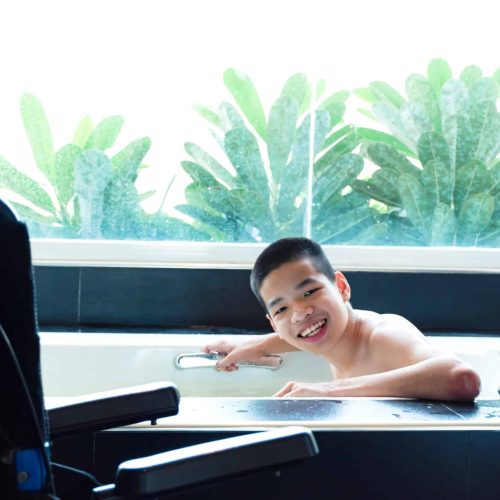 Hotels-Banner-scaled.jpg A smiling young man takes a bath at a spa with his wheelchair close by.