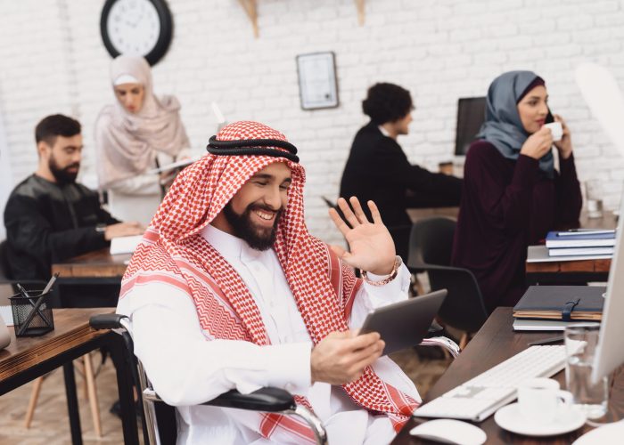 A disabled Arab man in a wheelchair holds a tablet in an office environment, he is smiling and waving to someone on a video call. In the background, a mixed group of adults work on computers, take sips of tea, talk to each other and on the phone to clients.