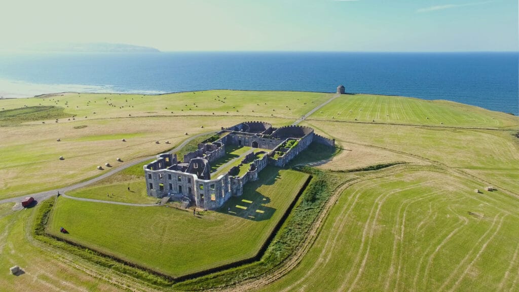 A photo of Mussenden Temple and Downhill Demesne taken from a helicopter. The castle grounds can be seen from the back and is surrounded on all side by well kept greenery. The temple sits in front of the castle further ahead on the edge of a cliff overseeing an untouched blue ocean.