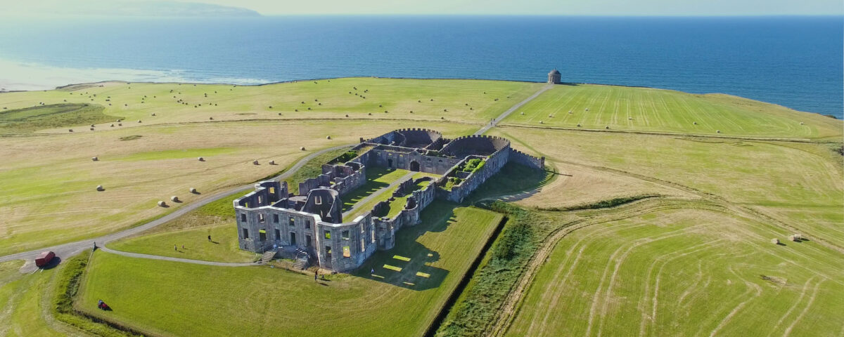 A photo of Mussenden Temple and Downhill Demesne taken from a helicopter. The castle grounds can be seen from the back and is surrounded on all side by well kept greenery. The temple sits in front of the castle further ahead on the edge of a cliff overseeing an untouched blue ocean.