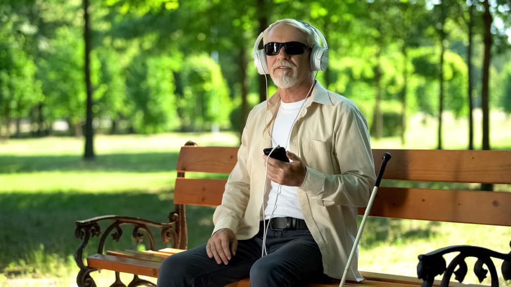 An old blind man on a park bench listens to an audio description on his phone with his cane leaning against the bench.
