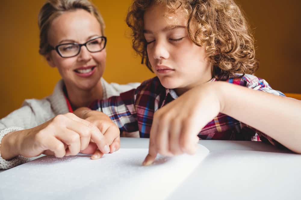 A young boy with eyes closed interprets braille with the help of a middle aged woman, who helps guide his hands.