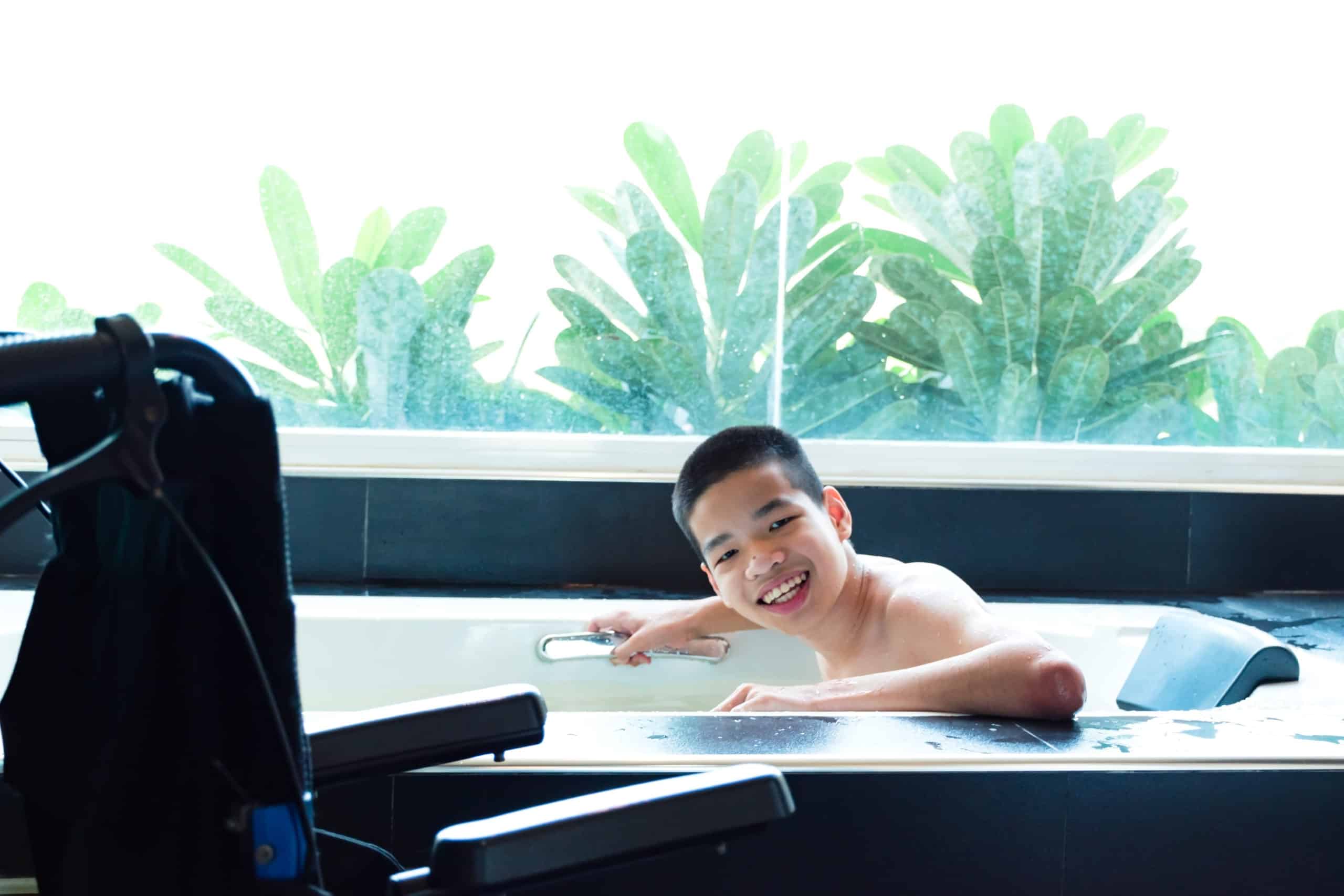 A smiling young man takes a bath at a spa with his wheelchair close by.