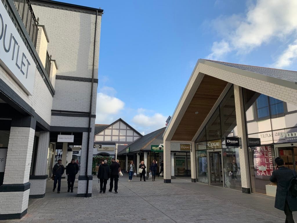 A photograph of Cheshire Oaks retail outlet on a sunny afternoon. A few cloud dot the otherwise blue sky as shoppers walk along the promenade. To the right of the image are Dune and Clogau stores.