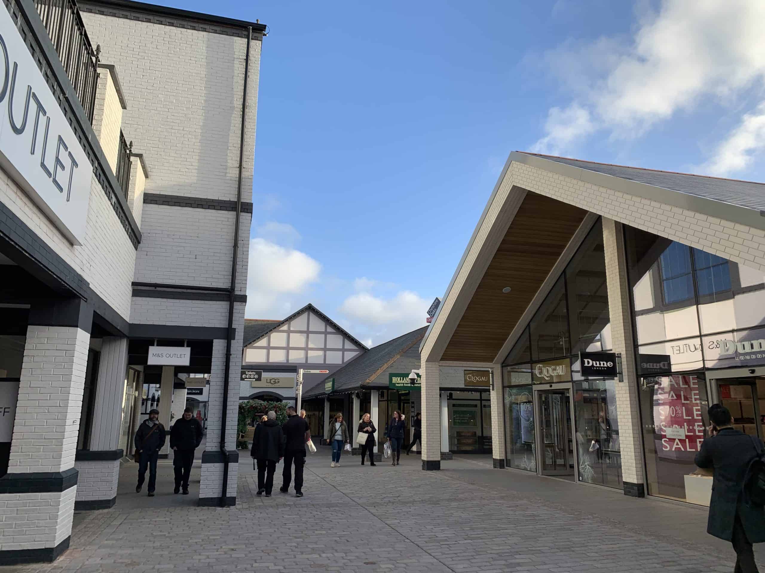 A photograph of Cheshire Oaks retail outlet on a sunny afternoon. A few cloud dot the otherwise blue sky as shoppers walk along the promenade. To the right of the image are Dune and Clogau stores.