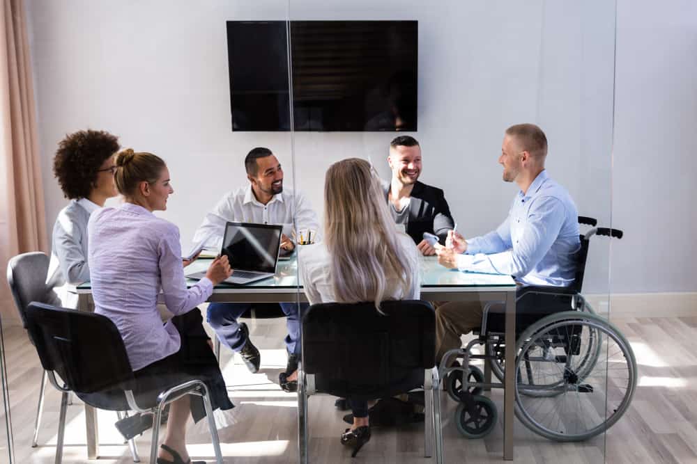 A mixed team of workers in a meeting in a conference room with a television in the background. One of the team is a man in a wheelchair.