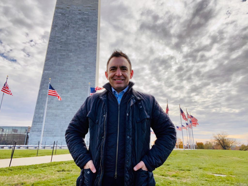 Steven Mifsud MBE poses for a photo with a field in the background with numerous U.S flags waving in the wind.