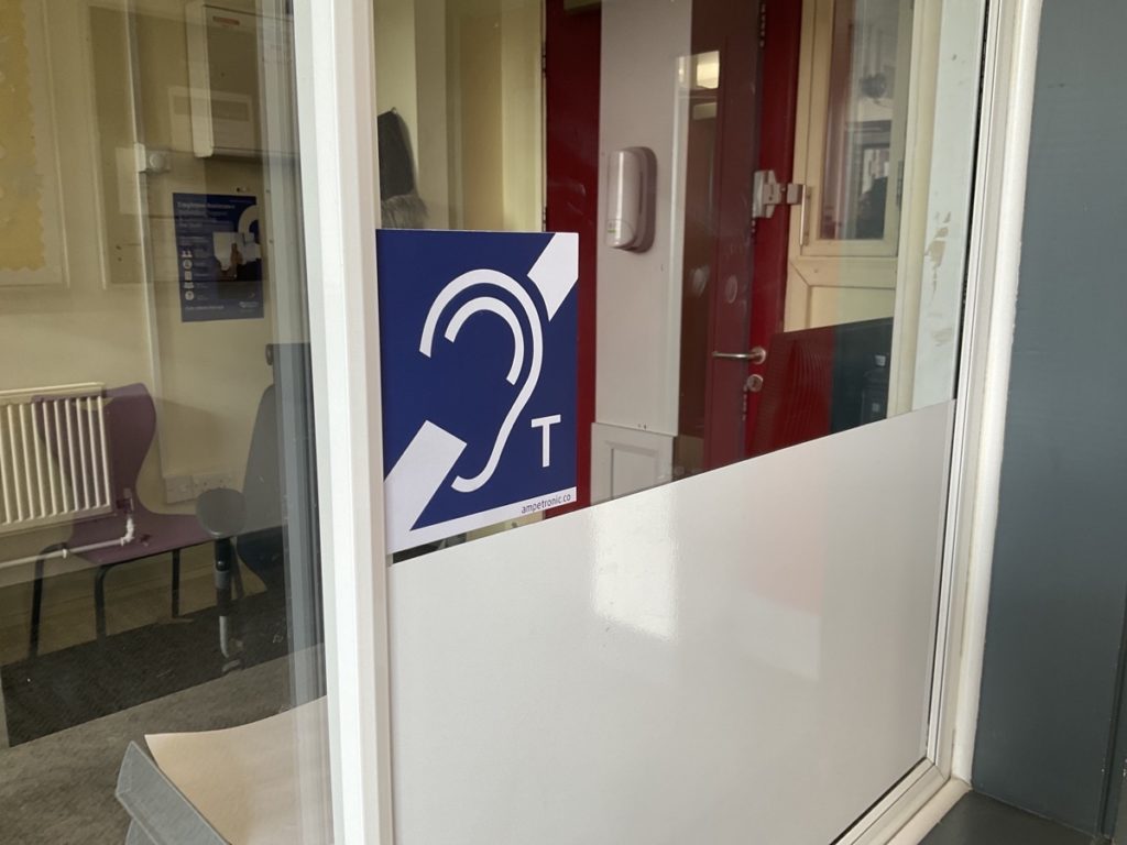 A Hearing Loop sign on a glass barrier outside a classroom.