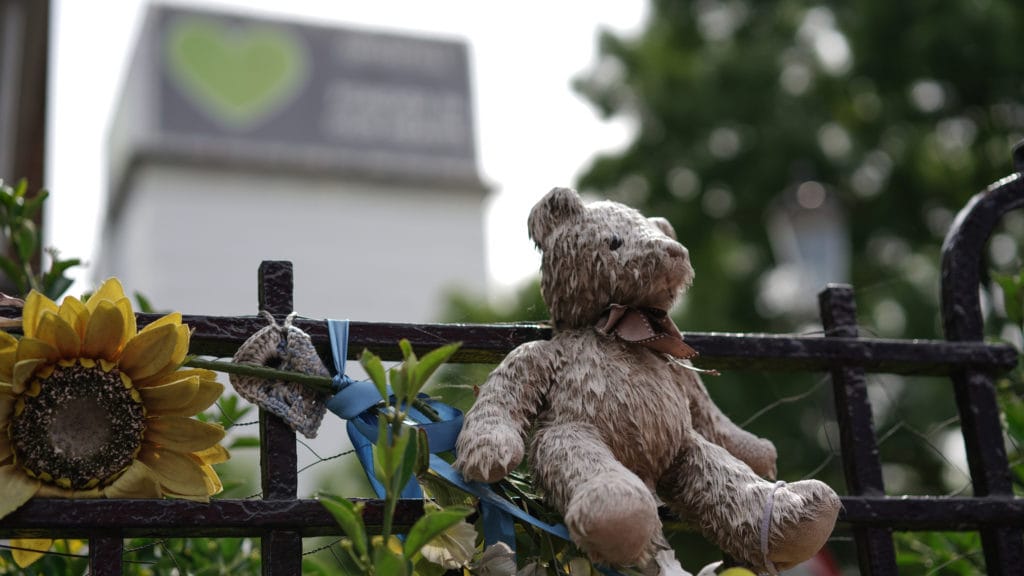 Teddy bear sat on a gate alongside other memorial items with Grenfell Tower London in the background
