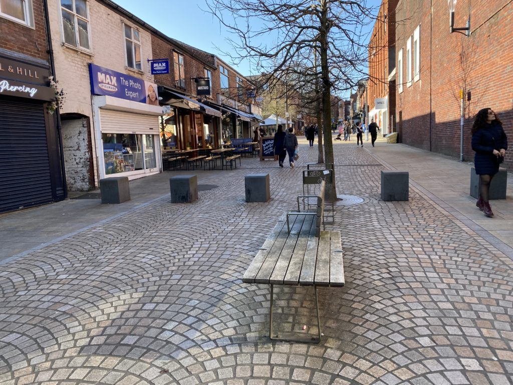 Altrincham town centre during the day. Several benches and cafes with seating outside. Shops include MAX Photo Experts and Marks and Spencers.