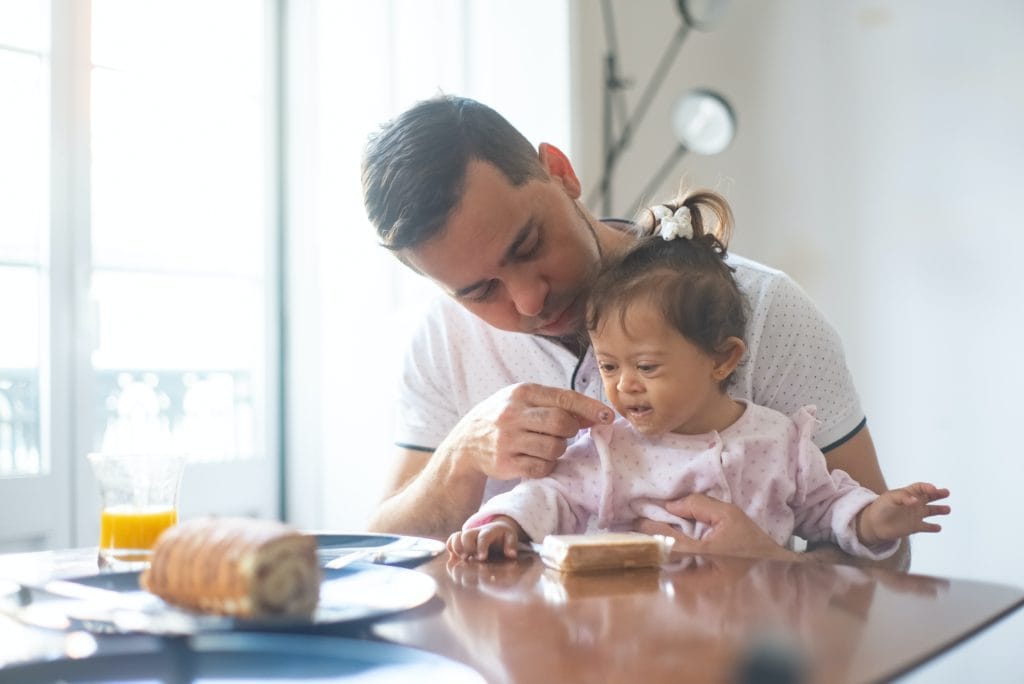 A father feeding his young disabled daughter some baby food at a table in their family home