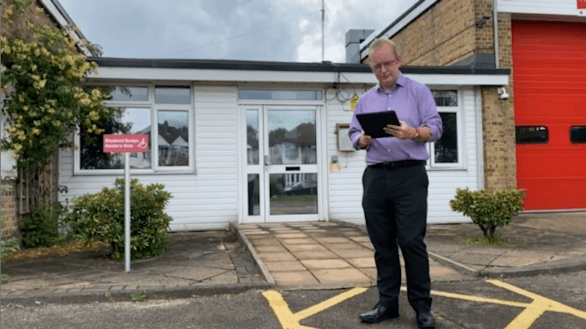 A male access consultant wearing smart clothes stands outside a fire station entrance holding and using a tablet