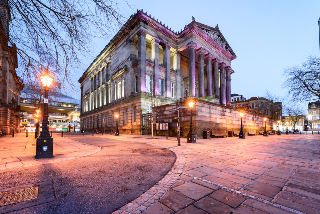 The Harris Museum Art Gallery Exterior, a grand imposing building supported by Greek style pillars from the front. Street lamps illuminate the front of the street below as the evening draws in.