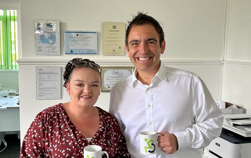A male and female member of the Direct Access team pose for a photo holding company branded mugs of tea and smiling at the camera in an office space. The mugs feature a photo of the wheelchair logo of Direct Access.