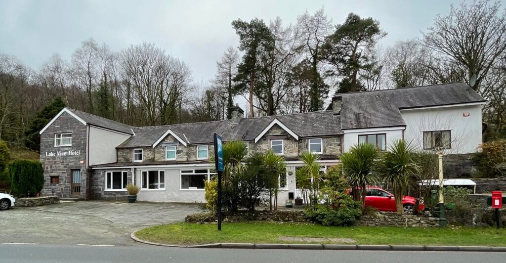 A photograph of the exterior of the Lake View Hotel, a long, white two story building situated on the edge of a road on an overcast day. Behind the site are some woods.