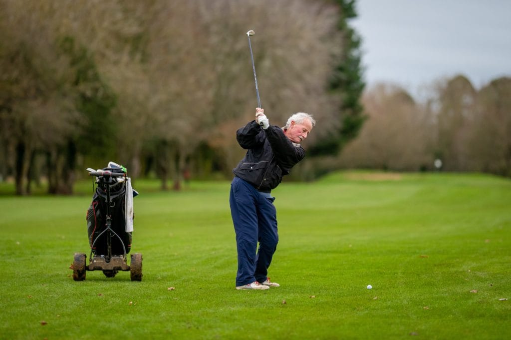 An elderly man on a green holding a golf driver lifts his arms backward and prepares for a shot