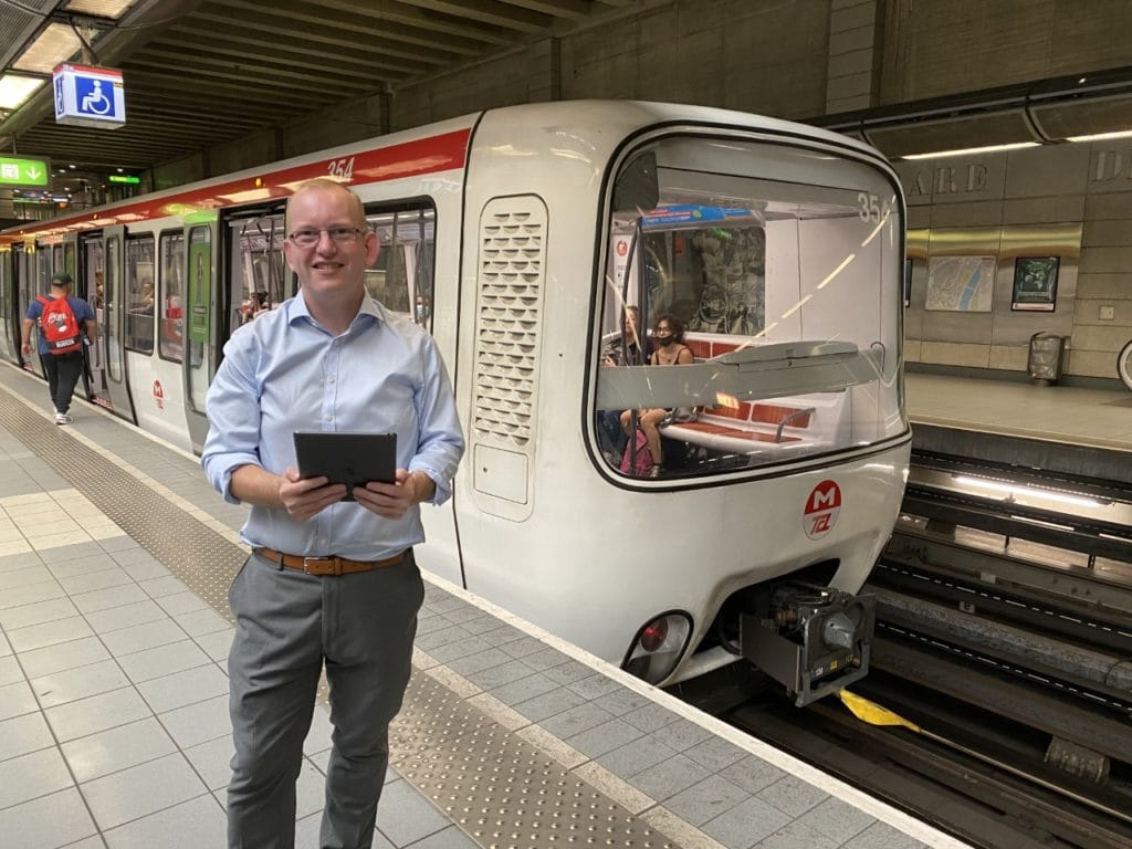 Middle aged man holding a tablet poses for a photo with a train on an indoor train platform.