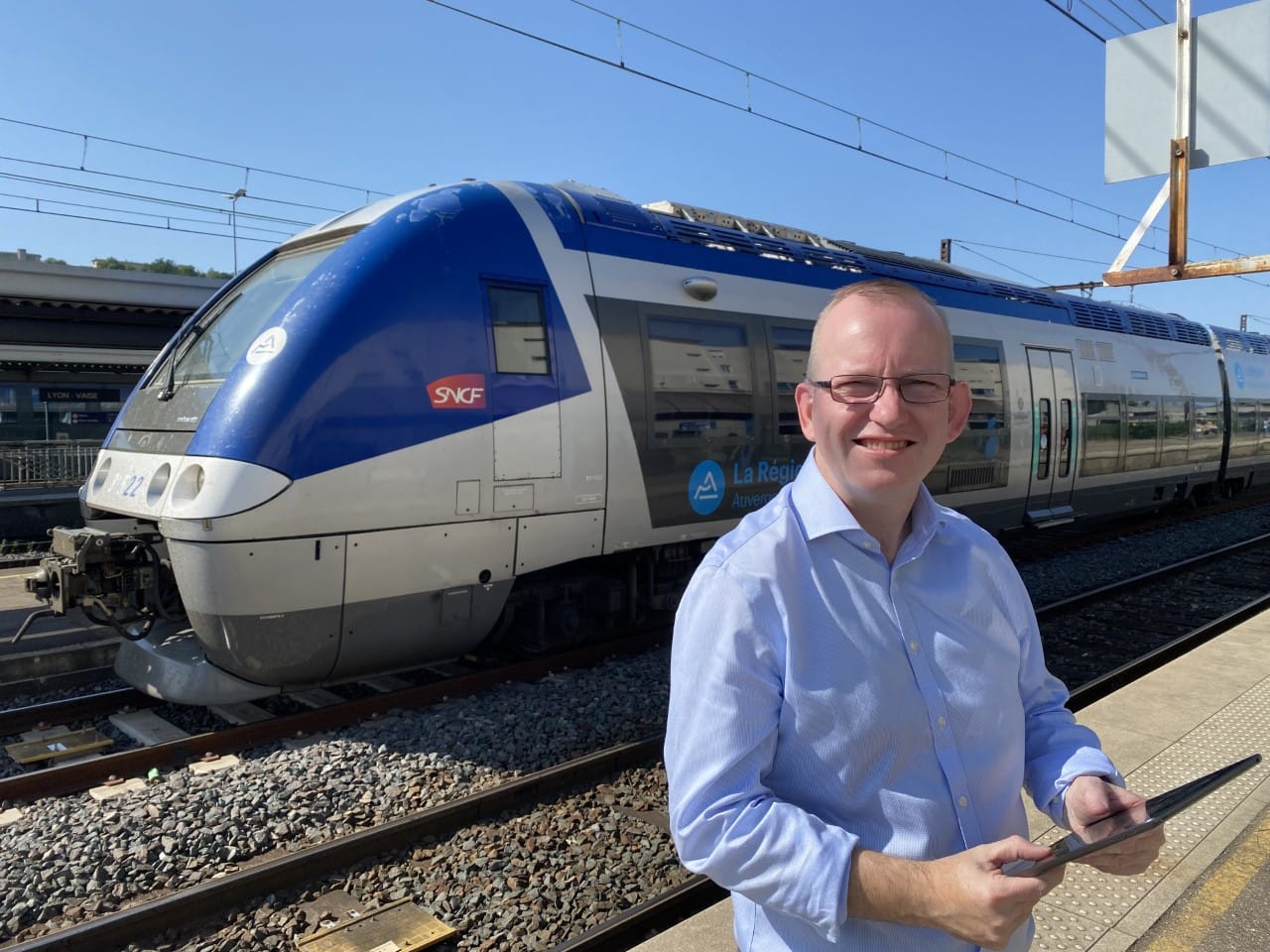 Middle aged man holding a tablet poses for a close-up photo with a train at Lyon station