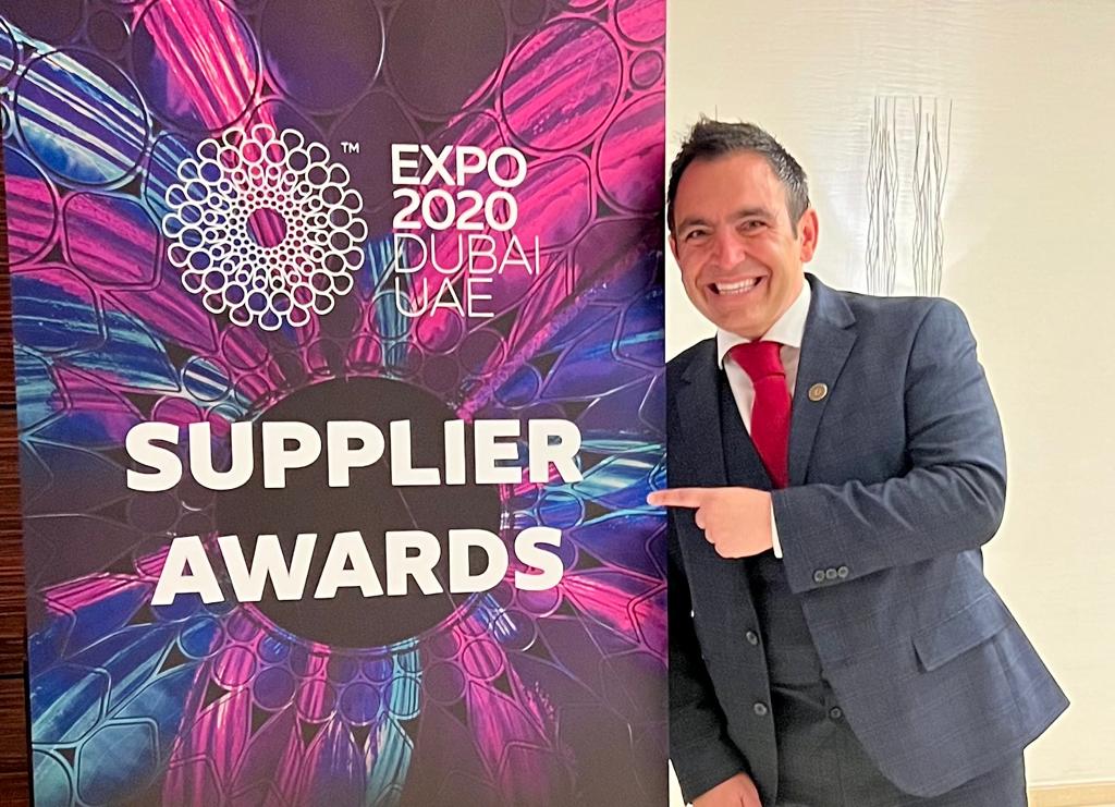 Middle aged man in a suit smiles pointing at an Expo 2020 Dubai Supplier Awards banner