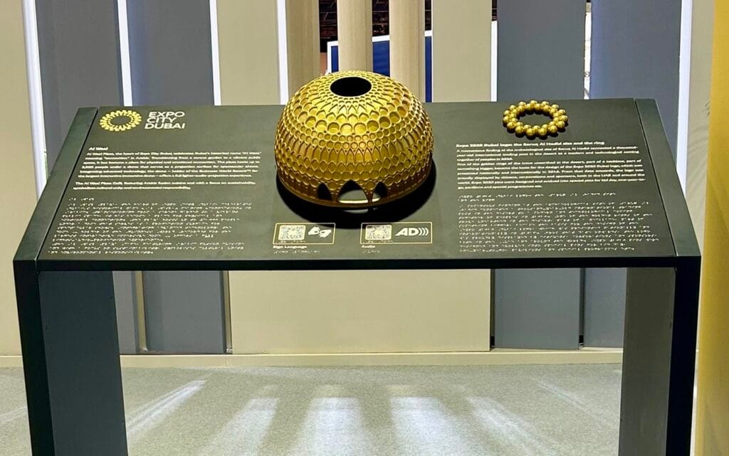 A shallow focus photo of a Direct Access tactile Map Board with a bustling conference in the background featuring various company exhibition stands and visitors walking around.