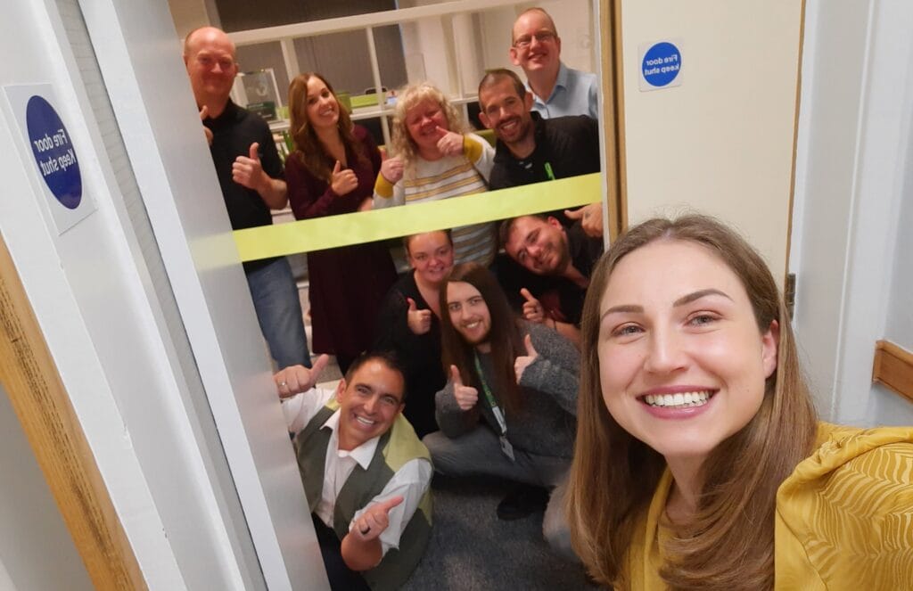 A group photo of the full Direct Access Team beaming smiles at the grand opening of the office. A yellow banner ready to be cut is stretched across the entrance.
