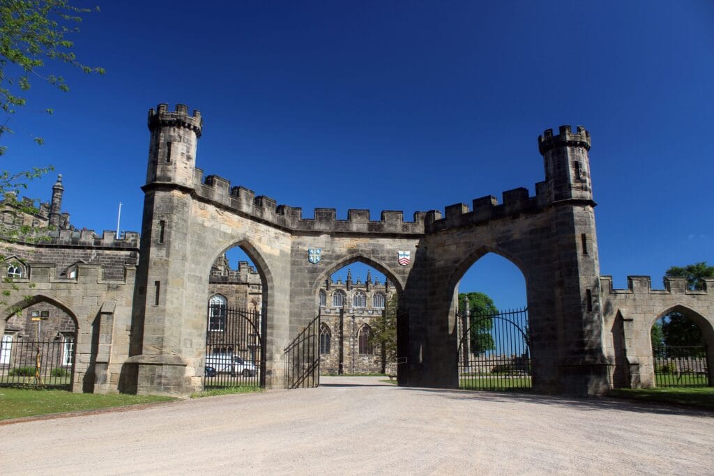 Auckland Castle's entrance seen from the exterior on a clear, sunny day. Three large gothic/baroque gates stand next to each other with the central one wide open. Behind the gate, a white car can be seen approaching the castle itself.