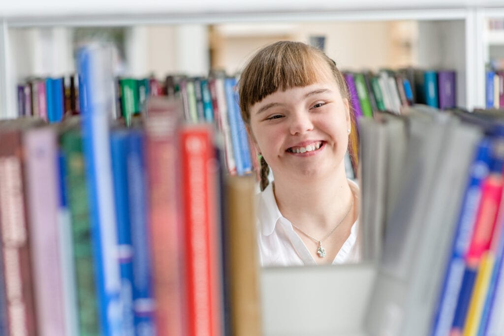 A smiling young schoolgirl with down syndrome chooses a book on a shelf in a brightly lit school library.