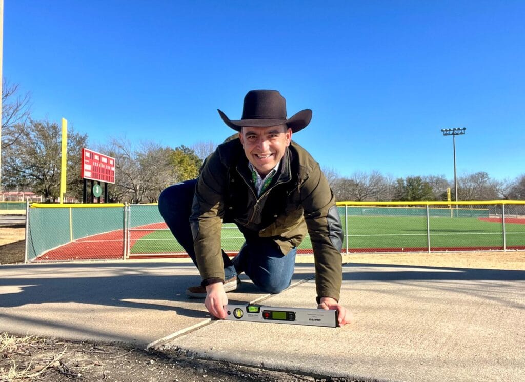 Direct Access founder Steven Mifsud MBE measuring the level of a concrete pathway with a baseball field in the background.