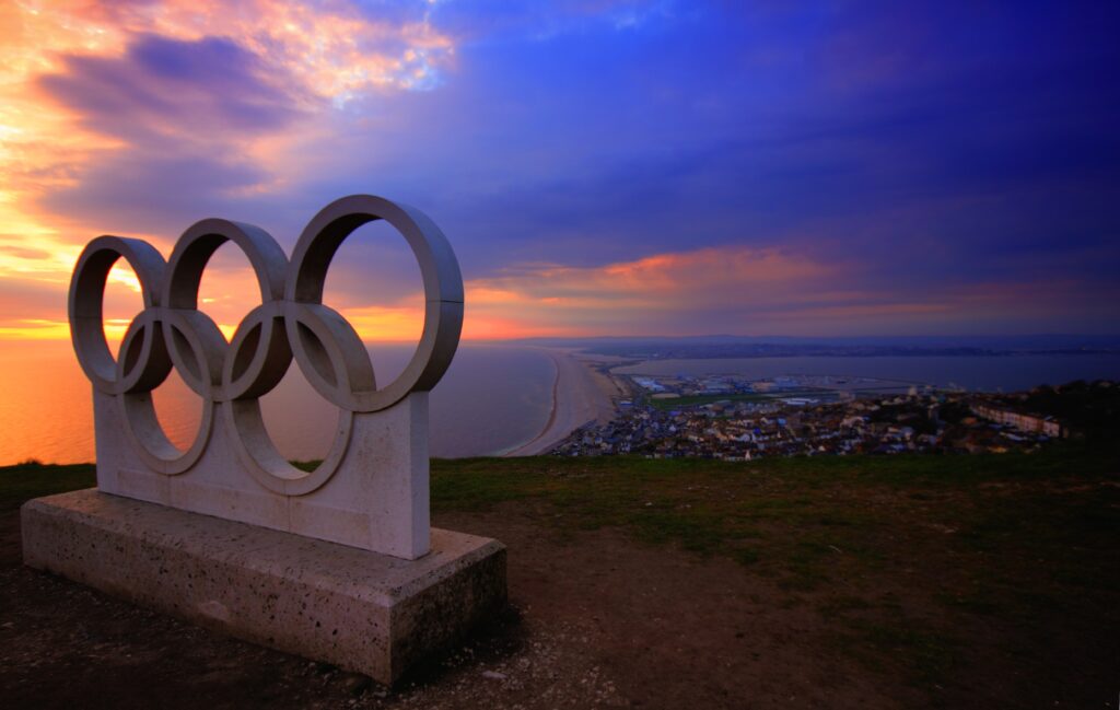 A photo of a large statue carved in the shape of the Olympics logo on top of a hillside at sunset. In the background is a town and the sea.