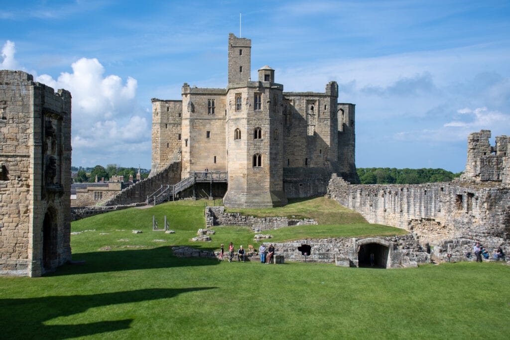Panoramic wide angle view of Warkworth Castle in Northumberland with tourists sat on a stone bench in the foreground.