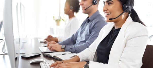 A smiling woman in a call center typing at her computer, speaking to a customer on a headset. Next to her are two colleagues, male and female, doing the same thing.