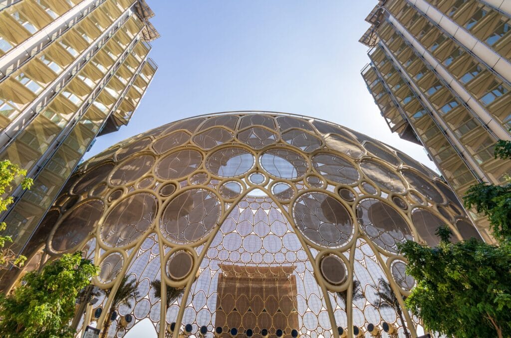 A large circular architectural structure with an intricate pattern of interlocking rings and translucent panels stands between two modern glass high‑rise buildings. The dome‑like design has a honeycomb appearance that lets light filter through. Trees at the bottom of the frame add greenery to the futuristic urban setting.