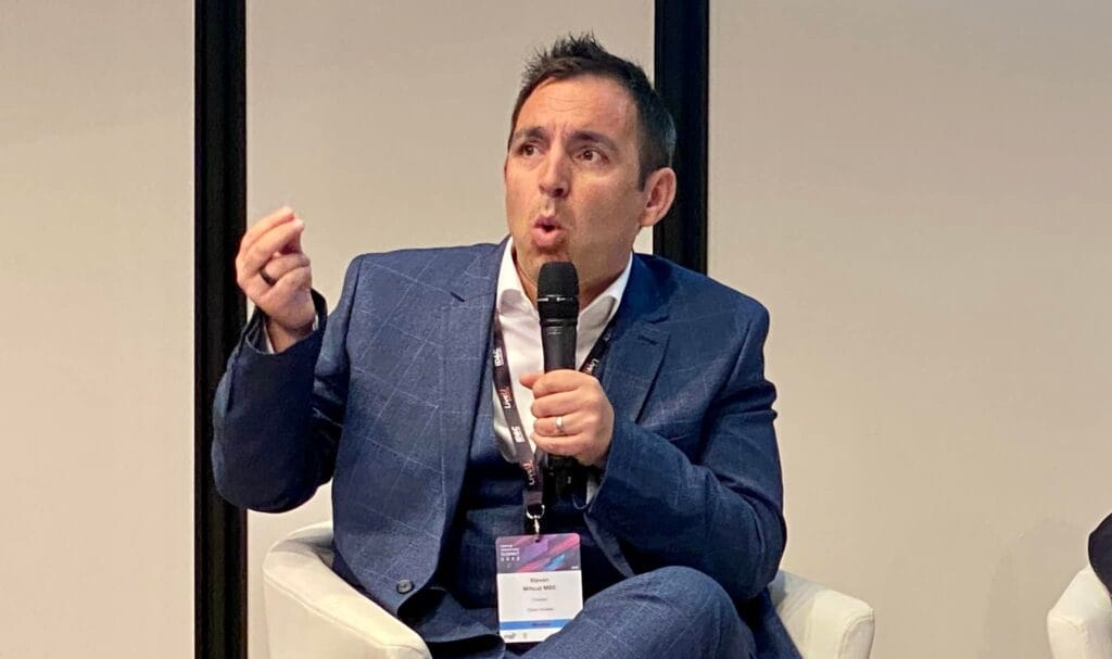 Steven Mifsud sitting in an armchair speaking at a conference holding his right hand in the air as if to make a point.
