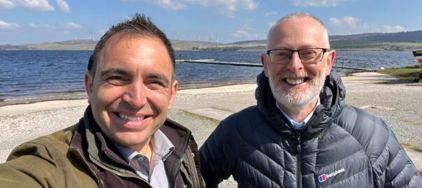 A selfie photograph of Steven Mifsud (Direct Access CEO) and Jamie Watson (Access Consultant) smiling with a lake and pier in the background on a cloudy day.