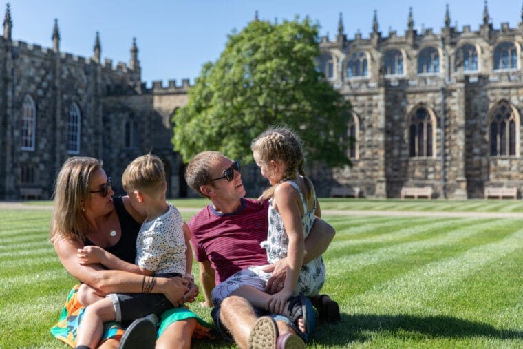 A man and woman wearing sunglasses sit on a freshly mown lawn on a sunny day with the backdrop of a castle in the background. Their children, a boy and a girl, sit on their laps.