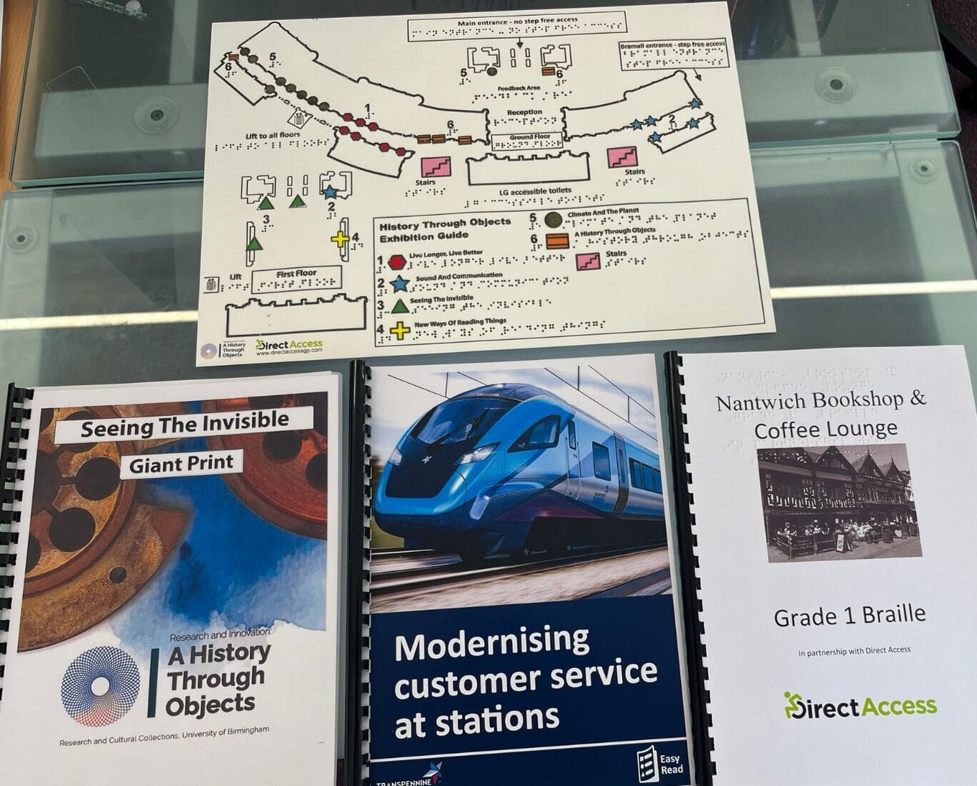 A selection of Direct Access' accessible media products laid out on a glass table next to each other. The products include a Giant Print guide, an Easy Read interpretation of a public information document, and a Braille translation of a coffee shop menu.