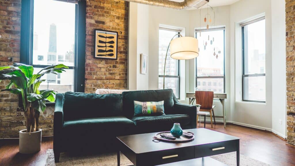 A tidy high-rise apartment living room with a green sofa, brown coffee table, small work desk near the windows and a large potted plant gently lit by the sun outside.