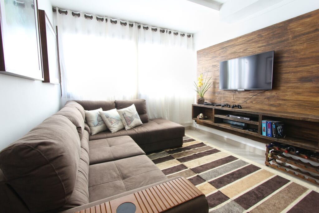 A wide-view of a modern living room with the sun permeating from behind white blinds. A brown corner sofa positioned in front a flat screen television. A counter below it has PlayStation remotes, games consoles, and film books.