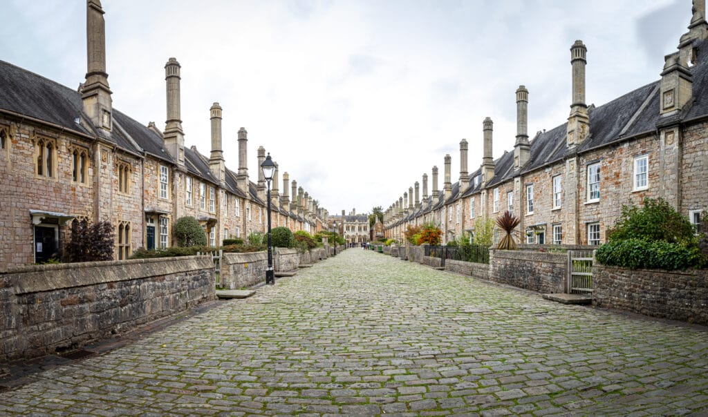 A photograph of Vicars Close in Wells, Somerset - a beautiful cobblestoned street with old historic houses lined up to the left and right sides. Beautiful gardens with variously coloured trees and plans are in front of each house.