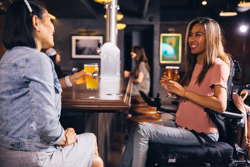 An Asian woman in a wheelchair enjoys a pint of beer with a friend in a pub.