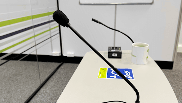 An induction loop microphone on a grey desk in an office environment. The photo also features a coffee mug and a black, rectangular hearing loop system.