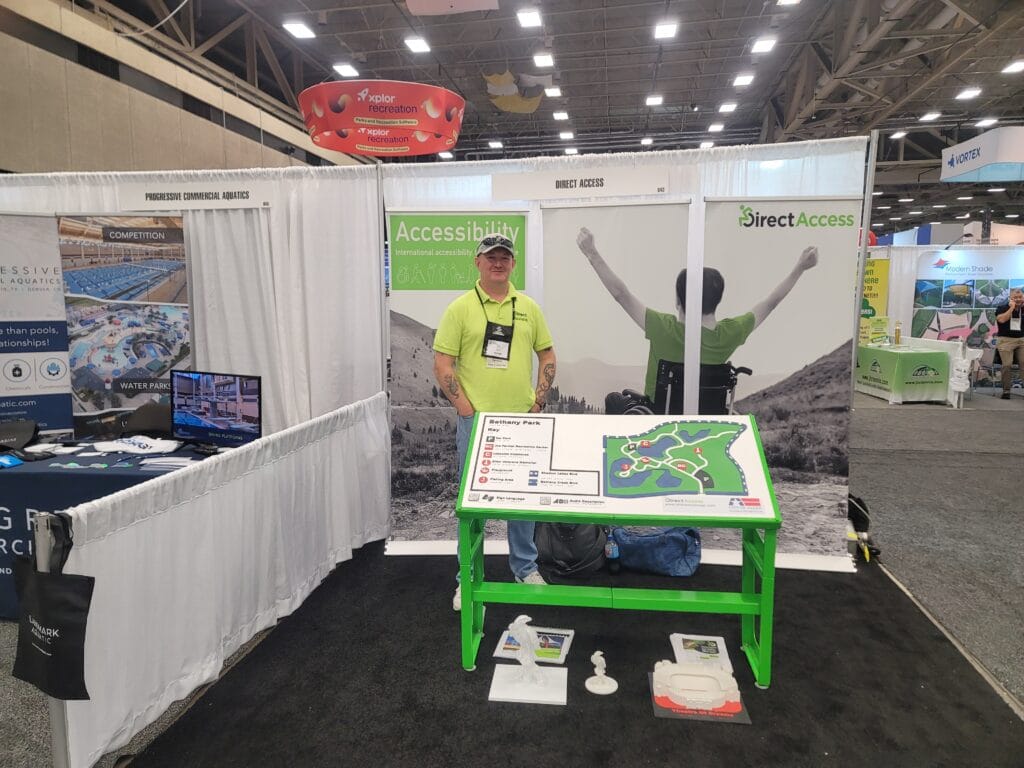 A middle aged man, Andy Lomas, poses for a photo behind a large tactile map board at a product and service conference. In the background is a poster banner for Direct Access showing a young man in a wheelchair cheering on a hillside.