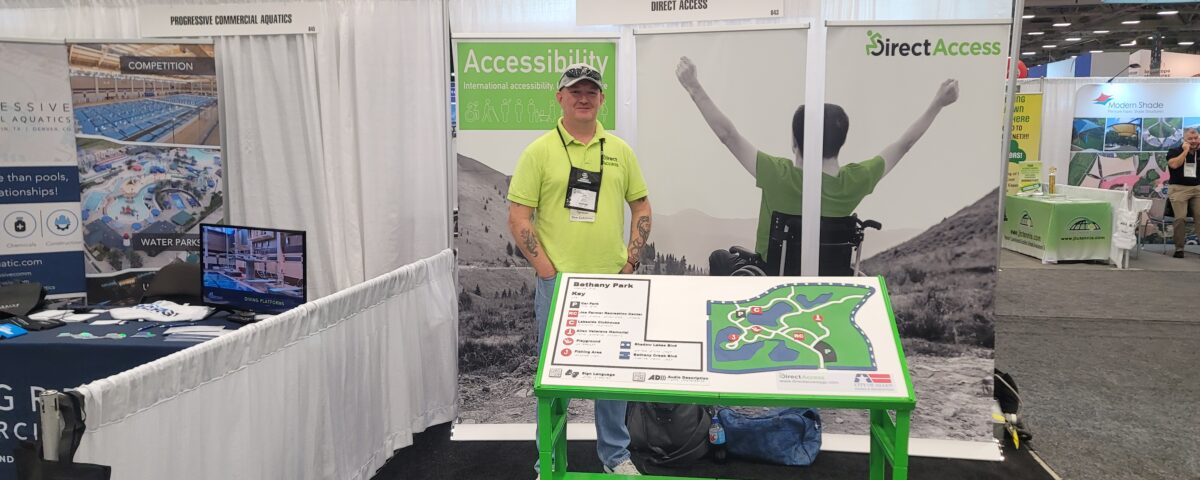A middle aged man, Andy Lomas, poses for a photo behind a large tactile map board at a product and service conference. In the background is a poster banner for Direct Access showing a young man in a wheelchair cheering on a hillside.