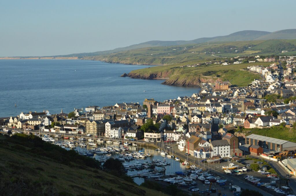 A panoramic view of an Isle of Man harbour and town during sunset. In the background are rolling hills and large fields.