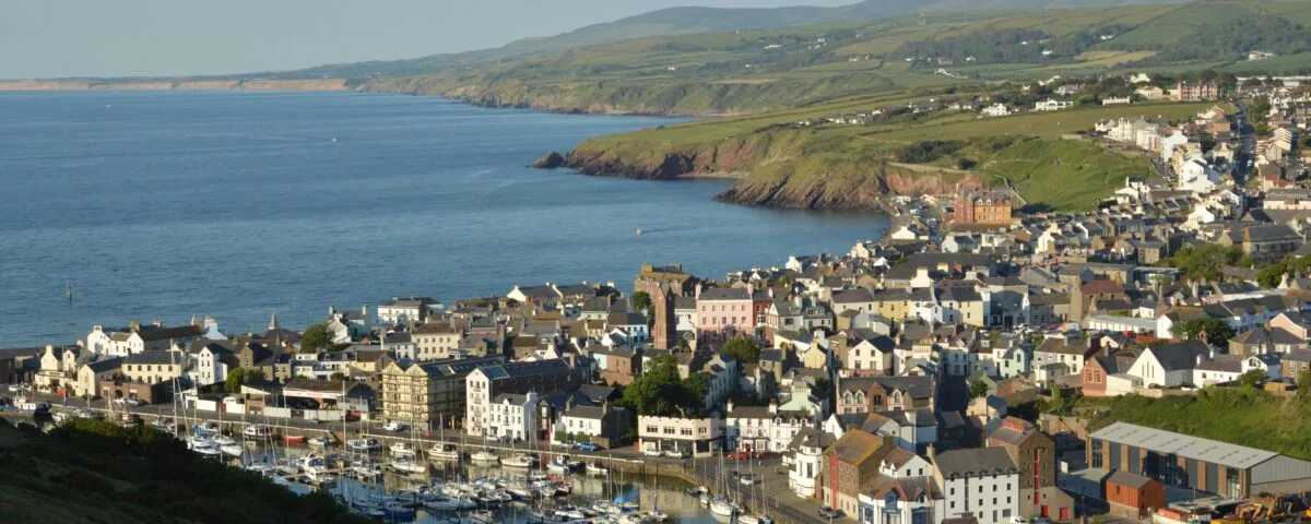 pexels-david-pryke-16709931 A panoramic view of an Isle of Man harbour and town during sunset. In the background are rolling hills and large fields.