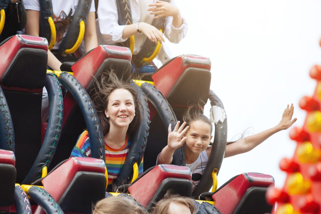 Two female children smile as a rollercoaster sends them flying down the track. One of them is posing for the camera holding her hands up high.