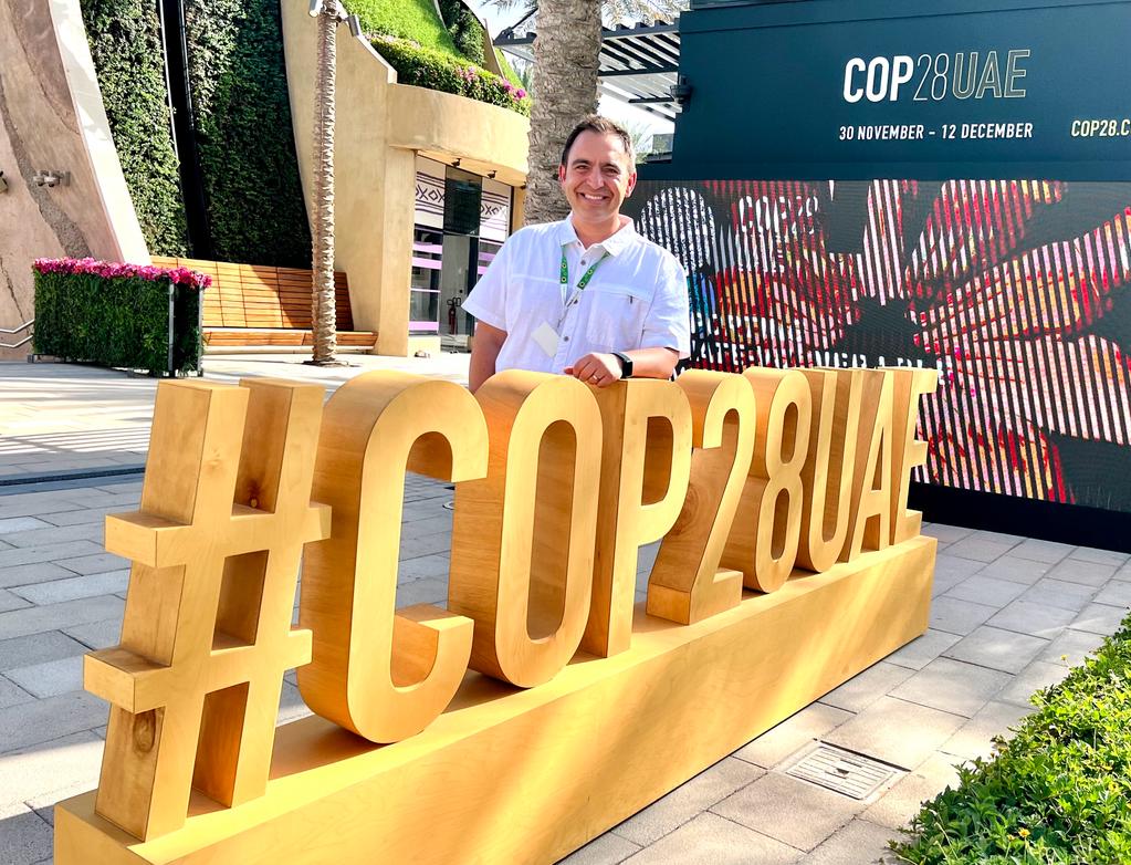 A photograph of a middle aged man wearing a white polo shirt and a hidden disability sunflower lanyard smiles and poses for a picture. His arm is resting on a large structure which reads #COP28UAE
